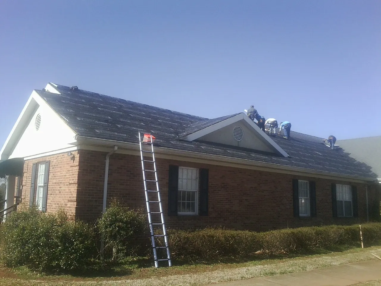Crew tearing shingles off a brick ranch roof with a ladder set — Emergency Roof Repair work by our crew in Luverne